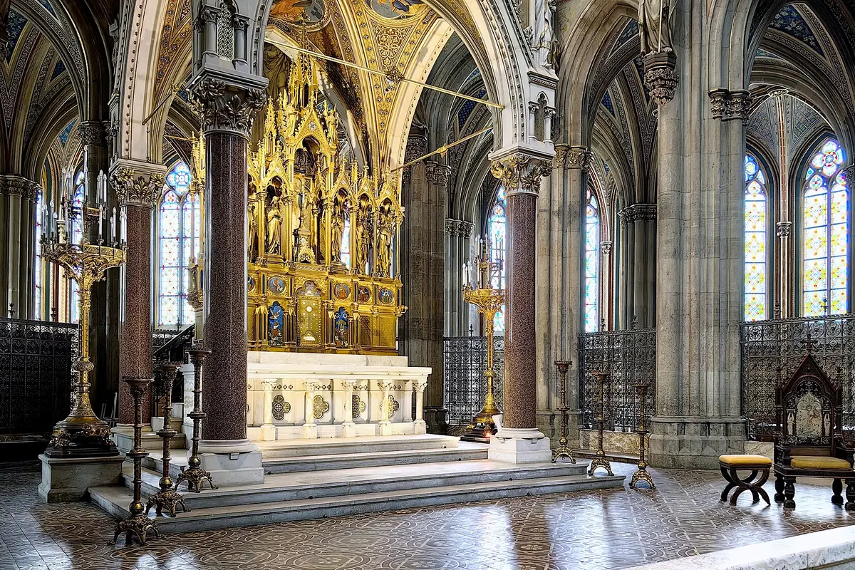 The gilded high altar of the Votivkirche on marble steps, framed by red granite columns, with a canopy of carved gold and stained-glass windows behind.