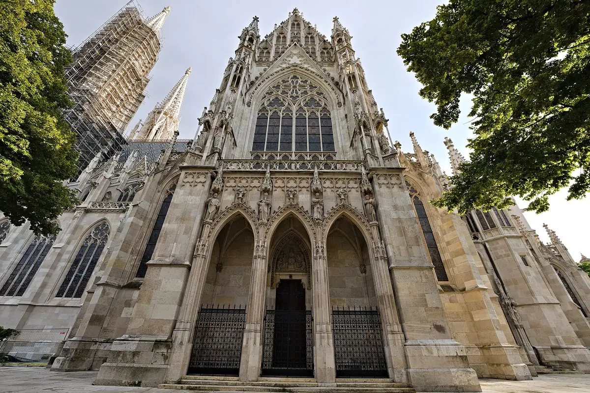The main portal of the Votivkirche, three pointed arched doorways under a large traceried window, with scaffolding on one tower at the left.