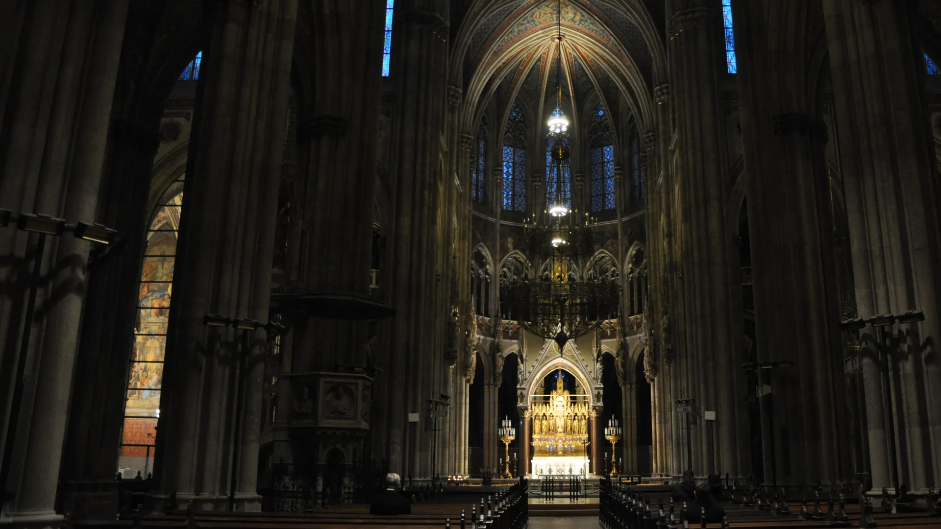 The nave of the Votivkirche in ordinary light: a dark aisle between tall grey stone piers leads to a gilded altar, with blue stained glass and a painted vault high above.